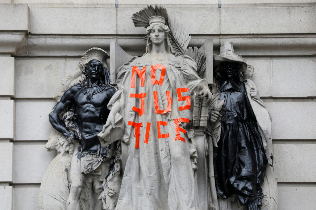 A Lady Justice statue outside New York City Hall with tape letters that read, "NO JUSTICE." Adjacent statues to the left and right are covered in black paint.