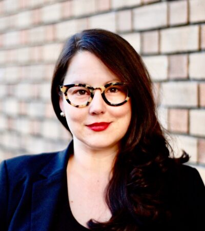 photo of person with dark hair, multicolor glasses and black top against a brick background