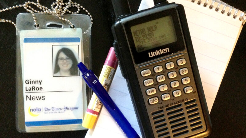 A photo of a press pass, reporter's notebook, a pen, and a police radio scanner.