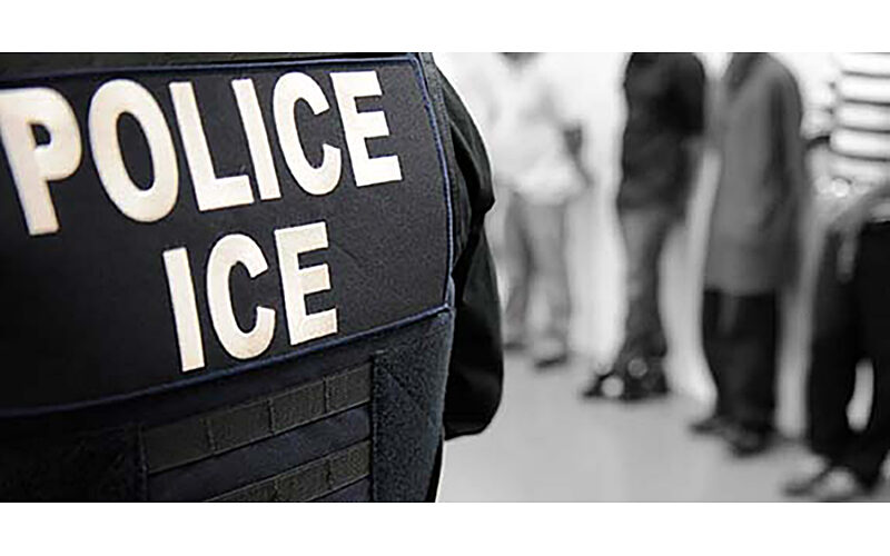 photograph of the back of an ICE officer's vest, with people standing in the background in a line.