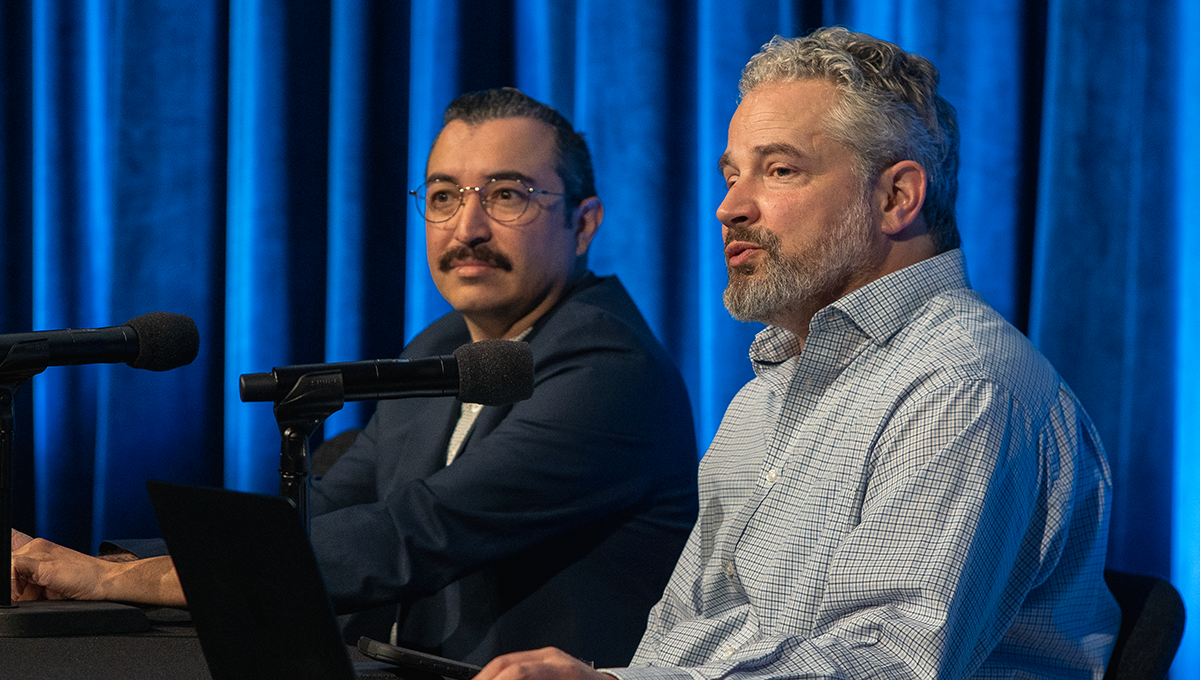 Two men seated at a table speaking before the audience.