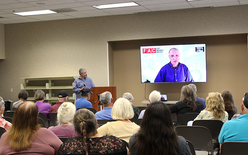 A meeting room with a man at the front and another man on a TV screen.